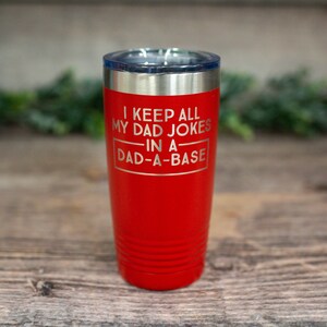 May include: A red insulated tumbler with a silver rim and a clear lid. The tumbler has the phrase "I KEEP ALL MY DAD JOKES IN A DAD-A-BASE" etched on the front. The tumbler is cylindrical with horizontal grooves near the bottom.