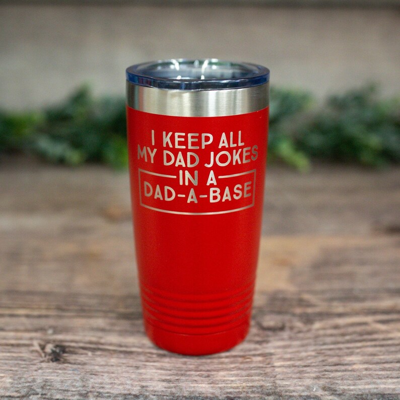 May include: A red insulated tumbler with a silver rim and a clear lid. The tumbler has the phrase "I KEEP ALL MY DAD JOKES IN A DAD-A-BASE" etched on the front. The tumbler is cylindrical with horizontal grooves near the bottom.