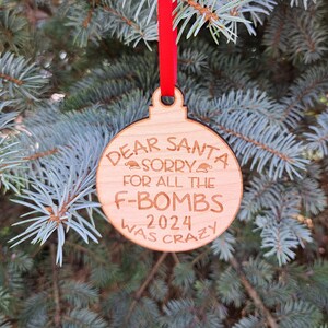 May include: A round wooden Christmas ornament with the text "Dear Santa Sorry for all the F-Bombs 2024 Was Crazy" on it. The ornament is hanging from a red ribbon.