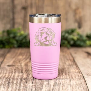 May include: A pink stainless steel tumbler with a white dog silhouette engraved on the front. The tumbler has a silver lid and a textured bottom.