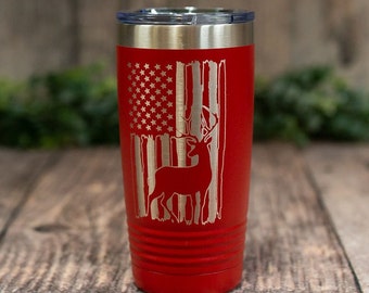 Puede incluir: Un vaso de acero inoxidable rojo con un diseño de bandera estadounidense desgastado y una silueta de ciervo. El vaso tiene una tapa y está aislado.