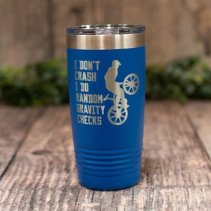 May include: A blue stainless steel tumbler with a silver lid and a silver inscription that reads "I don't crash I do random gravity checks" with a silhouette of a person riding a bicycle.