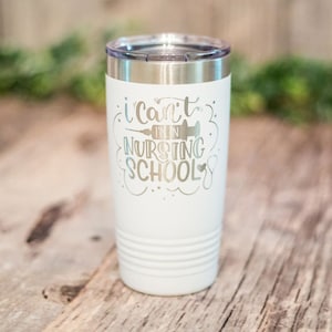 Op de afbeelding: Een witte geïsoleerde beker met een zilveren deksel. De beker heeft de tekst "I can't I'm in Nursing School" in zilveren letters, met een spuit- en stethoscoopafbeelding. De beker heeft een geribbeld ontwerp aan de onderkant.