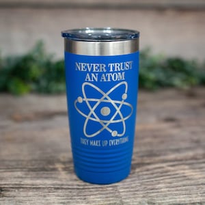 May include: A blue insulated tumbler with a silver lid. The tumbler features the text "NEVER TRUST AN ATOM" above an atom symbol, and "THEY MAKE UP EVERYTHING" below. The tumbler is designed for cold and hot beverages.