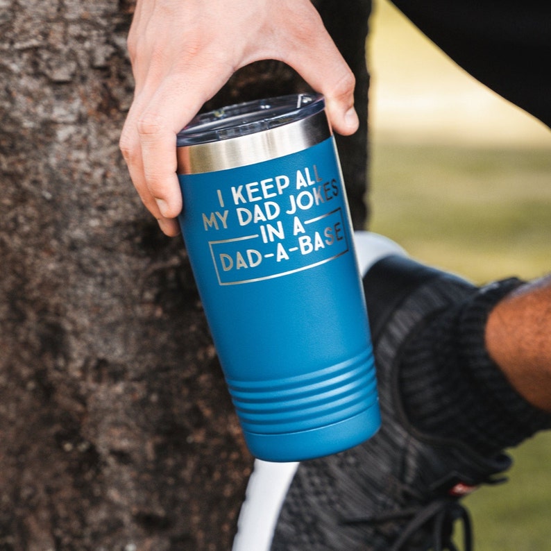 May include: A blue insulated tumbler with a silver rim and lid. The tumbler has the text "I KEEP ALL MY DAD JOKES IN A DAD-A-BASE" in silver lettering. The tumbler is held by a person.