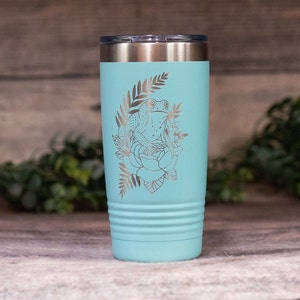 May include: A teal stainless steel tumbler with a silver engraved design of a frog sitting on a flower with leaves.