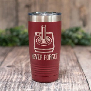 May include: A red, stainless steel tumbler with a white engraved image of a vintage video game joystick and the text "NEVER FORGET".
