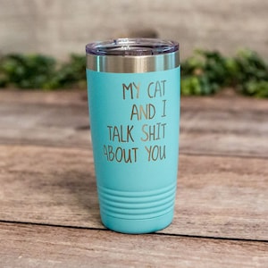 May include: A light blue insulated tumbler with a silver lid. The tumbler has the text "MY CAT AND I TALK SHIT ABOUT YOU" in gold lettering. The tumbler is cylindrical with a ridged bottom.