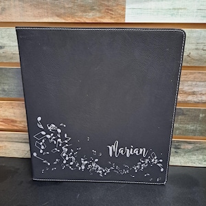 May include: Black leather binder with a white stitched border. The binder has a design of musical notes and the name "Marian" engraved on the front.