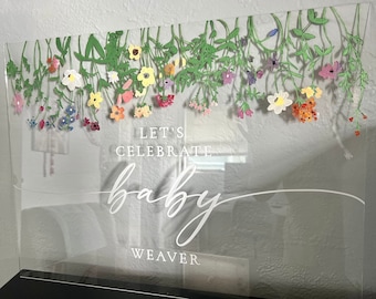 May include: A clear acrylic sign with a floral design and the text "Let's Celebrate Baby Weaver".