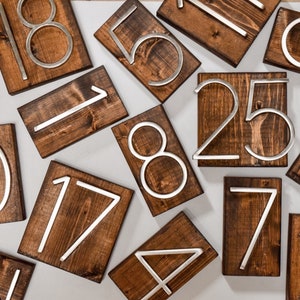 Puede incluir: Un conjunto de placas de n&uacute;meros de madera con n&uacute;meros de metal plateado. Los n&uacute;meros est&aacute;n dispuestos en un patr&oacute;n aleatorio sobre una superficie blanca.