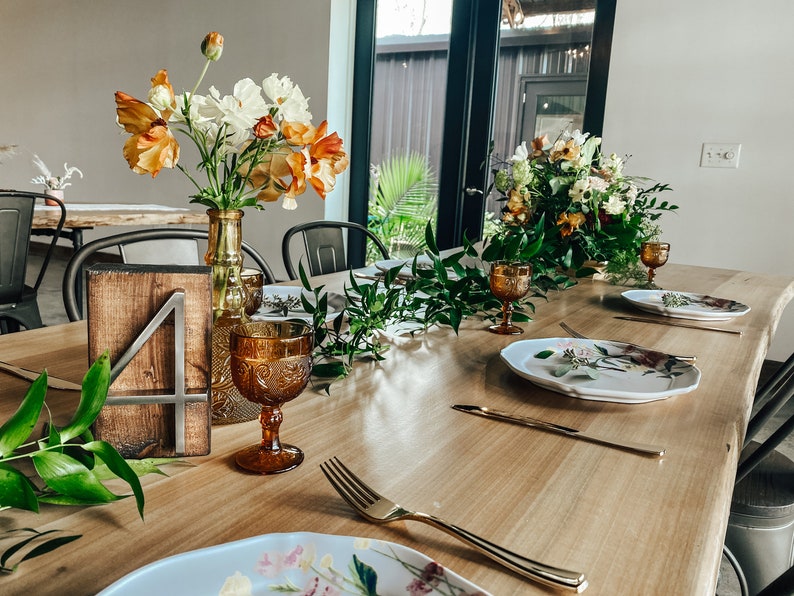 Peut inclure: Une table en bois dress&eacute;e pour une occasion sp&eacute;ciale, avec des compositions florales, de la verrerie ambr&eacute;e et des assiettes d&eacute;coratives. Un num&eacute;ro de table en bois 4 est visible. La table est dress&eacute;e avec des couverts dor&eacute;s et de la verdure.