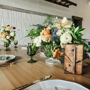 Peut inclure: Une table en bois dress&eacute;e pour une r&eacute;ception, avec des centres de table floraux dans des vases en verre, des gobelets verts et des assiettes avec des couverts dor&eacute;s. Un num&eacute;ro de table en bois avec le chiffre 1 est visible.