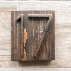 Puede incluir: Un letrero de madera con el n&uacute;mero 17 en letras de metal plateado. El letrero es de color marr&oacute;n oscuro y tiene un aspecto r&uacute;stico.