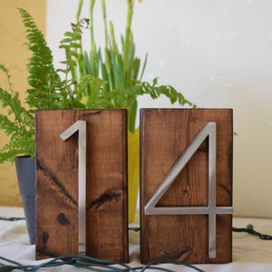 Peut inclure: Deux blocs de num&eacute;ros de table en bois, l'un avec un "1" en m&eacute;tal et l'autre avec un "4" en m&eacute;tal. Les blocs sont de couleur marron fonc&eacute;. Les chiffres sont argent&eacute;s. L'arri&egrave;re-plan comprend des plantes et des guirlandes lumineuses.