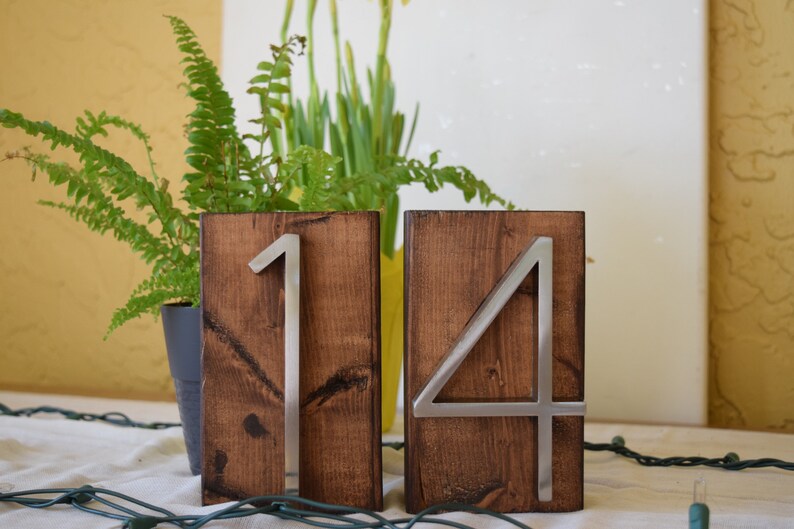 Peut inclure: Deux blocs de num&eacute;ros de table en bois, l'un avec un "1" en m&eacute;tal et l'autre avec un "4" en m&eacute;tal. Les blocs sont de couleur marron fonc&eacute;. Les chiffres sont argent&eacute;s. L'arri&egrave;re-plan comprend des plantes et des guirlandes lumineuses.