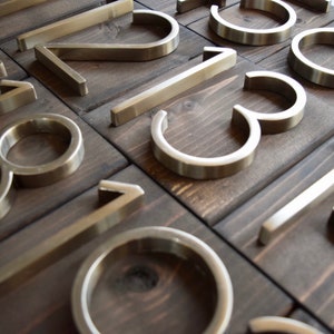 Puede incluir: Un conjunto de n&uacute;meros de casa de metal plateado sobre un fondo de madera oscura. Los n&uacute;meros son modernos y elegantes, con un acabado cepillado.