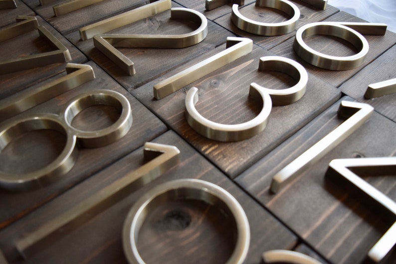 Puede incluir: Un conjunto de n&uacute;meros de casa de metal plateado sobre un fondo de madera oscura. Los n&uacute;meros son modernos y elegantes, con un acabado cepillado.