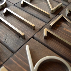 Puede incluir: Primer plano de n&uacute;meros de casa de metal plateado sobre una superficie de madera marr&oacute;n oscuro. Los n&uacute;meros son modernos y elegantes, con un dise&ntilde;o plano y minimalista.