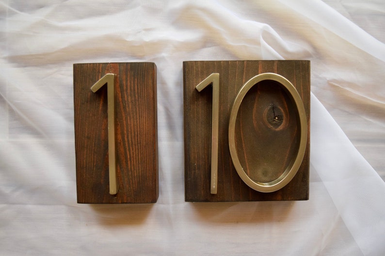 Peut inclure: Deux blocs de bois avec les chiffres "1" et "10" fix&eacute;s. Les chiffres sont m&eacute;talliques et mont&eacute;s sur du bois teint&eacute; fonc&eacute;. Le "0" est de forme ovale. Le bois a un grain visible.