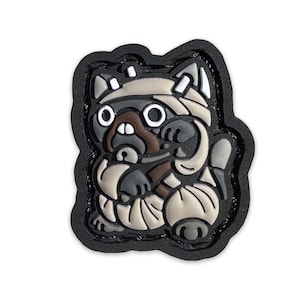May include: A black and white cartoon pug wearing a white and brown mummy costume. The pug has a surprised expression on its face. The patch is made of rubber and has a black border.