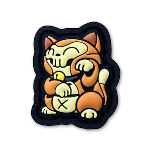 Puede incluir: Un parche de goma negro y naranja con una imagen de dibujos animados de un gato sonriente con una cara de calavera. El gato lleva un collar con una campana y tiene una moneda de oro en su pata.