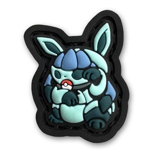 Pode incluir: Um patch de desenho animado colorido com uma criatura com detalhes em azul, verde-azulado e preto. O design inclui uma bola vermelha e branca, em um fundo preto. O patch tem um efeito 3D.