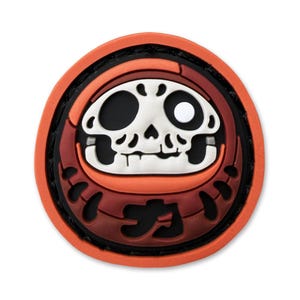 May include: A circular patch in shades of orange, red, and black, with a skull design. The skull has one black eye and one white eye. Japanese characters are at the bottom of the patch.