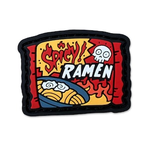 May include: A rectangular rubber patch featuring a cartoon graphic of a bowl of noodles with the text "SPICY! RAMEN" in red and white. The design includes a white skull icon, orange flames, and a blue bowl on a red and black background.
