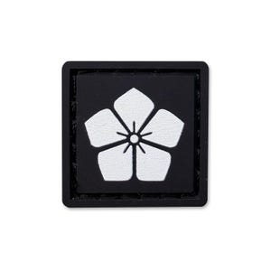 May include: A square patch featuring a black background with a white five-petaled flower emblem centred on the surface. The design has a raised, textured appearance with a thick black border framing the central floral graphic element.