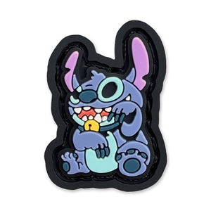 May include: A blue and purple Stitch character from the Disney movie Lilo & Stitch. The character is sitting with its arms crossed and has a mischievous grin. The character is made of a soft, textured material and is attached to a black patch.