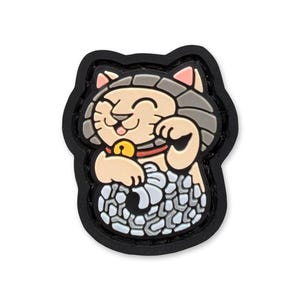 Könnte beinhalten: Ein lächelnder, hellbrauner Maneki-neko-Katzenanhänger mit erhobener Pfote, rotem Halsband und goldener Glocke. Die Katze ist vor einem schwarzen Rand platziert, mit einer grau-weißen gemusterten Basis.