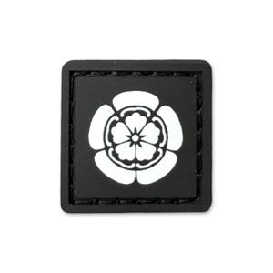 May include: A square, black patch with a white floral crest design. The crest is centrally placed and has a detailed, symmetrical appearance. The patch has a stitched border.