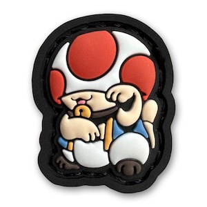 May include: A red and white mushroom character with a pacifier in its mouth, wearing a blue and white outfit. The character is sitting with its arms crossed and has a black outline.