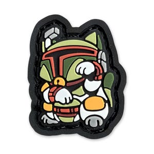 Op de afbeelding: Een zwarte en groene geborduurde patch met een cartoon afbeelding van Boba Fett uit Star Wars. De patch is katvormig en heeft een wit gezicht met een rode mond en gele ogen.