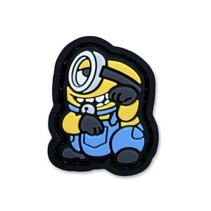 May include: A rubberized shoe charm featuring a yellow cartoon character with a single large silver-rimmed eye, wearing blue overalls and black gloves. The character has a wide grin showing white teeth and is outlined in a thick black border.