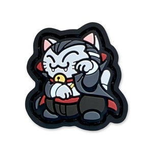 May include: A black, white, gray, and red Maneki Neko cat charm in a vampire costume. The charm has a black border and features a smiling cat with a cape, collar, and bell.