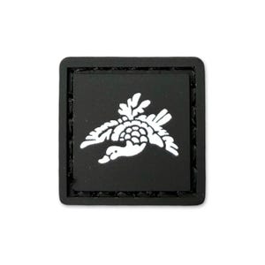 May include: A square, black patch featuring a white duck in flight. The duck is depicted with detailed wings and a textured body. The patch has a stitched border and is set against a plain white background.
