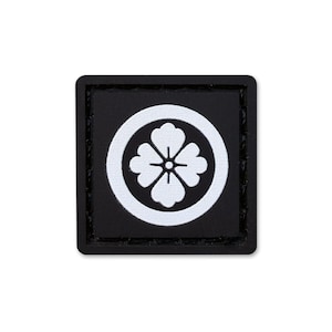 Puede incluir: Un parche cuadrado negro que presenta un borde circular blanco que encierra un emblema floral estilizado de cuatro pétalos en color blanco. El parche tiene un marco negro sólido con un borde texturizado de alto contraste.
