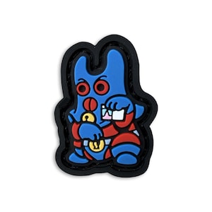 May include: A blue, red, and yellow rubberized charm featuring a stylized creature with two ears, red circular eyes, and a red nose. The figure holds a yellow key and a white rectangular object, outlined in black with a raised, textured surface.