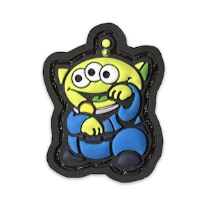 May include: A green alien character with three eyes, wearing a blue jumpsuit, is depicted on a black patch. The alien is smiling and has a playful expression.