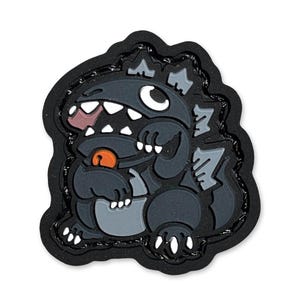 May include: A cartoon-style patch featuring a gray monster with white teeth, a pink tongue, and an orange ball. The monster is set against a black background with a textured border. The design is a playful and whimsical representation.