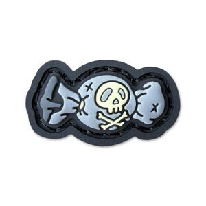 May include: A black and grey rubberized patch shaped like a wrapped candy. The center features a pale yellow skull and crossbones icon. The design is outlined in a thick black border, creating a distinct, raised, and durable appearance for gear.