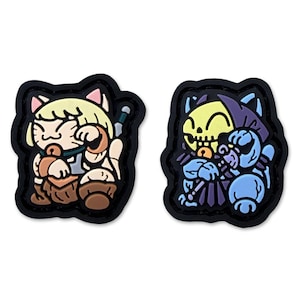 May include: Two rubberized shoe charms with black borders. The left charm features a cream-colored cat character with blonde hair and brown clothing. The right charm depicts a blue and purple skeletal cat character with a yellow skull face.
