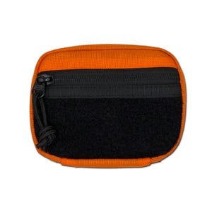May include: A rectangular orange pouch featuring a black zippered compartment and a large black hook-and-loop fastener patch on the front. The zipper includes a black cord pull tab, and the overall construction consists of durable woven fabric.