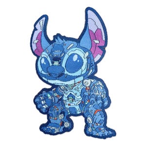 May include: A blue and white embroidered patch featuring a stylized robot-like Stitch character. The patch has a detailed design with shades of blue, pink, and orange, and is set against a white background. The character has large eyes and distinctive ears.