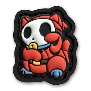 Pode incluir: Um patch de personagem de desenho animado vermelho, branco e preto. O personagem tem um rosto branco semelhante a um crânio com olhos grandes e pretos, um corpo vermelho e uma borda preta. O patch é um design 3D.