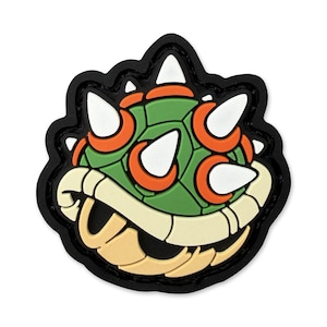 Peut inclure: Un écusson coloré représentant le visage d'un personnage de dessin animé. Le design comprend une carapace verte, des accents orange, des pointes blanches et une bouche beige, le tout souligné de noir. Pour vêtements ou accessoires.