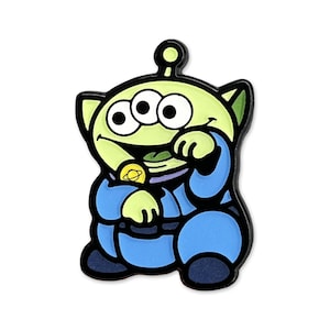 Può includere: Spilla con personaggio alieno verde con tre occhi, che indossa un'uniforme blu. La spilla ha un contorno nero e un emblema dorato sul petto. L'alieno sorride e tiene in mano un oggetto verde.