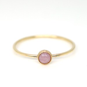 May include: A delicate gold ring featuring a small, round, light pink gemstone. The ring has a thin, polished gold band and a simple, elegant design. The gemstone is set in a bezel setting, adding a touch of color and sophistication.